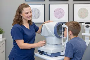 A young boy is having his eyes examined by a woman in a blue uniform in an office with framed prints on the wall