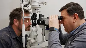 A man wearing a blue shirt and a ring is looking through an ophthalmoscope at another man who is wearing a gray shirt and a blue watch. The man in the gray shirt is looking at the man in the blue shirt.