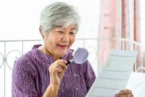 An older woman is examining a document through a magnifying glass, while sitting on a bed with a pink curtain in the background.