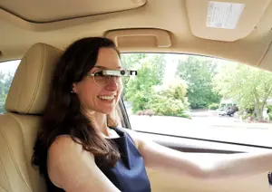 A woman wearing a blue sleeveless shirt is smiling while sitting in a car with a beige interior and a window showing a road with trees and a building.