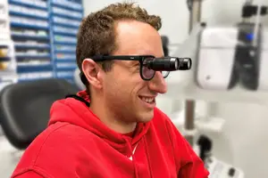A man with binoculars wearing a red hoodie smiling at the camera in a room with medical equipment