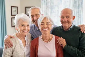 A group of four smiling elderly people posing for a photo in a room with a blue curtain and picture frames on the wall