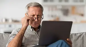 A man sitting on a couch and holding a laptop with one hand covering his eyes, probably tired.