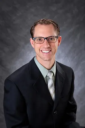 Professional headshot of a smiling man wearing glasses, a suit, and tie