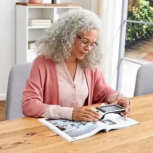 Older woman wearing glasses reads an open magazine at a table with a handheld device.