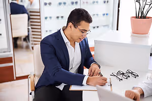 A man is sitting on a chair and writing on a paper while looking at a pair of eyeglasses.