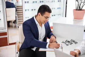 A man is sitting on a chair and writing on a paper while looking at a pair of eyeglasses.