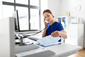 A woman in a blue shirt is sitting at a desk and talking on the phone