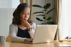 An Asian woman working on a laptop while sitting at a table in a room with a plant and a window in the background