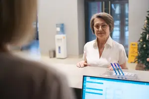 An older woman wearing glasses is smiling and looking at something on the right. She is probably inside an office. In front of her is a desk with a monitor, a bottle, and a pen. Behind her is a blurry person, a Christmas tree, a water dispenser, and a glass door. On the right is a yellow sign on the wall.