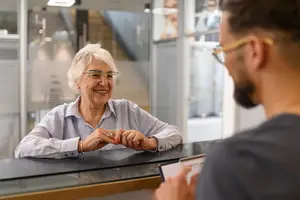 A smiling elderly woman in glasses is talking to a man in an office setting