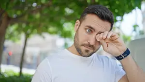 Man wearing white shirt and blue watch rubbing his eyes while looking to the side