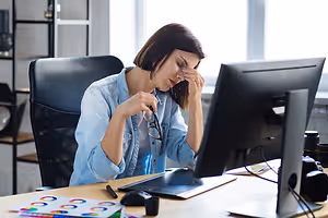 A woman is sitting on a chair in front of a desk. She has her eyes closed and is holding her head with her left hand. She is probably working on the computer. There is a monitor, a mouse, a pen, a pencil, a notepad, and a lamp on the desk. There are also some other things on the desk. Behind her, there is a glass wall with a view of the outside.
