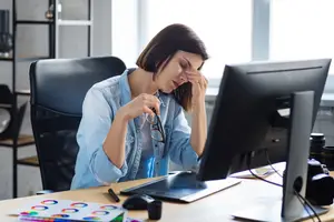 A woman is sitting on a chair in front of a desk. She has her eyes closed and is holding her head with her left hand. She is probably working on the computer. There is a monitor, a mouse, a pen, a pencil, a notepad, and a lamp on the desk. There are also some other things on the desk. Behind her, there is a glass wall with a view of the outside.