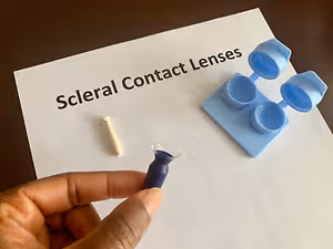 A person holds a scleral contact lens next to a lens case and lens holder on a white sheet of paper