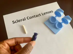 A person holds a scleral contact lens next to a lens case and lens holder on a white sheet of paper