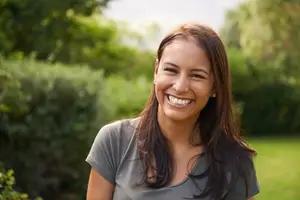 A smiling woman with brown hair wearing a gray t-shirt is standing in a garden with green grass and bushes.