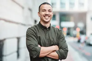 A smiling man with folded arms standing in front of a building on a city street