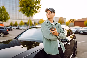 A man is standing beside a black car in a parking lot and holding a cell phone.
