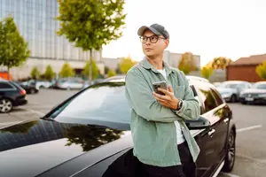 A man is standing beside a black car in a parking lot and holding a cell phone.