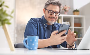 A man is sitting at a desk with a laptop and a cup, holding a cell phone in his hand, smiling, and looking at the screen.