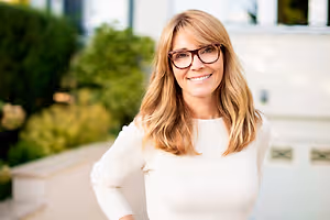 Portrait of a smiling woman with glasses and a white top standing in front of a house with plants