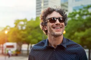 Man with curly hair wearing sunglasses smiling in front of a city street with trees and buildings