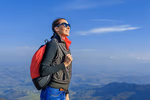 A woman wearing a backpack and a red and black jacket is standing on a mountain peak, smiling and looking at the view.