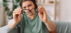 A woman wearing a green shirt is sitting on a couch and examining a pair of black glasses.
