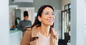 A smiling woman in glasses is standing in front of a white wall in a shop, with a blurred image of a woman standing in front of a desk in the background.