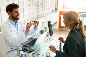 A man in a white lab coat and a woman in a black coat are sitting at a table with a laptop and a phone. The man is holding a pair of glasses and smiling while the woman is looking at him and smiling. Behind them is a glass display case with eyeglasses and a glass window with a reflection of a window.