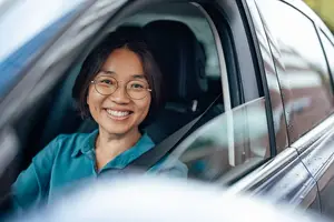 A smiling woman with glasses drives a car with her seat belt fastened
