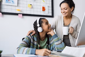 A woman with headphones and a cup in her hand is smiling while sitting in front of a desk with a young girl in a sweater, who is sitting with a smile on her face while looking at her laptop, with a woman standing behind her and smiling as well.