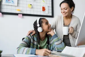 A woman with headphones and a cup in her hand is smiling while sitting in front of a desk with a young girl in a sweater, who is sitting with a smile on her face while looking at her laptop, with a woman standing behind her and smiling as well.
