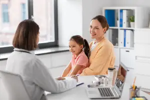 A doctor is talking to a girl and her mom in a room with a desk and a laptop