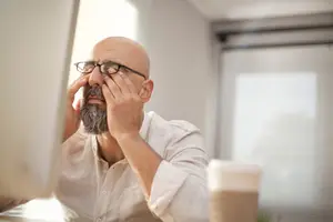 A man sitting in front of a desk with his hands on his face, wearing glasses and a white long-sleeve shirt