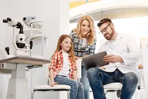 A young girl is sitting on a chair in an optometrist's office with her parents and a doctor examining her eyes using an ophthalmoscope