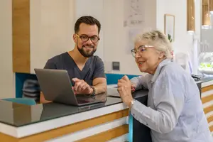 A man and an elderly woman are standing in a medical clinic and smiling while looking at the laptop