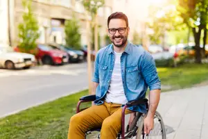 A smiling man with a disability in a wheelchair on a sidewalk beside a street with parked cars