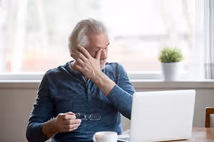 An older man with a headache sitting in front of a laptop