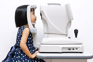 A young girl with short black hair wearing a blue dress with white polka dots is sitting on a chair and looking into an eye examination machine