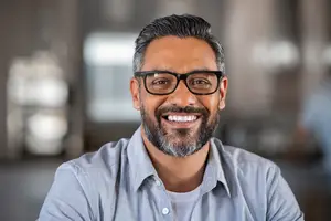 A man wearing glasses and a blue shirt is smiling and posing for a photo in an office room