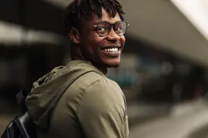 Portrait of a smiling man with glasses and a green hoodie carrying a backpack standing outside a building