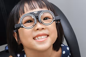 A young girl wearing glasses with a 54 written on the lens, she is smiling and looking at something in front of her, probably at an eye clinic.