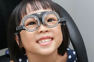 A young girl wearing glasses with a 54 written on the lens, she is smiling and looking at something in front of her, probably at an eye clinic.