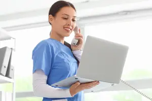 A woman in a blue scrub is holding a laptop and talking on a phone while smiling