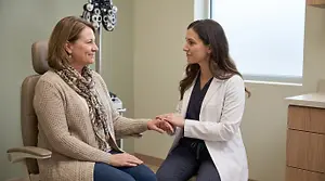 An older woman and a younger woman are sitting in chairs and talking to each other in a room with an eye exam machine and a window in the background.