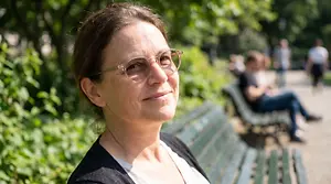 An adult woman wearing glasses is sitting on a park bench and smiling at the camera.