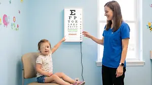 A young girl with a patch on her eye is reading an eye chart in a doctor’s office.