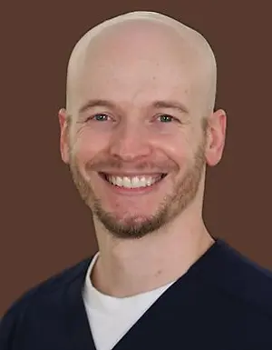 Portrait of a smiling man with a bald head and wearing a navy blue jacket over a white shirt against a brown background